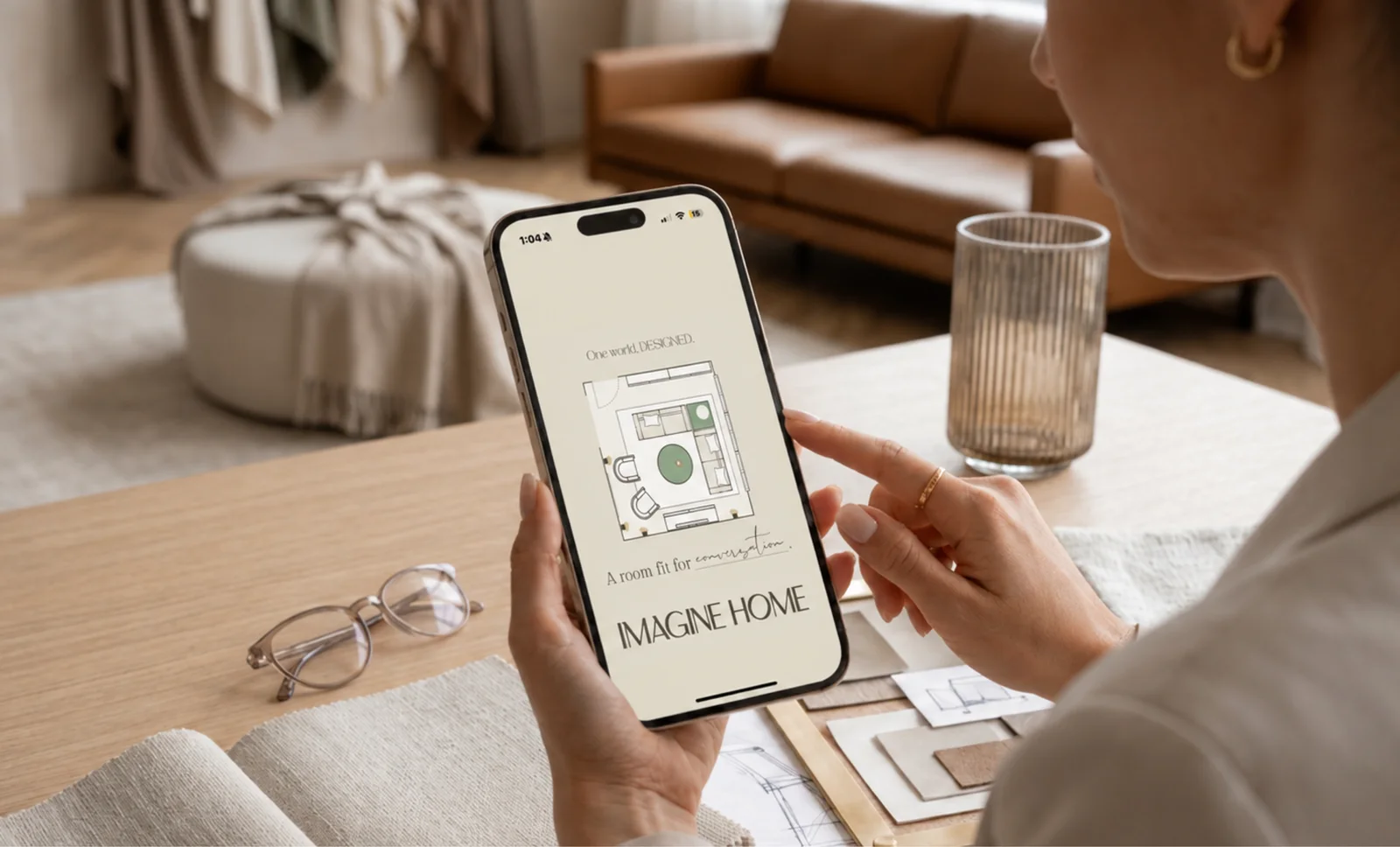 A designer holds a phone showing the Imagine Home app with a room floor plan, in a warm styled interior with material swatches.