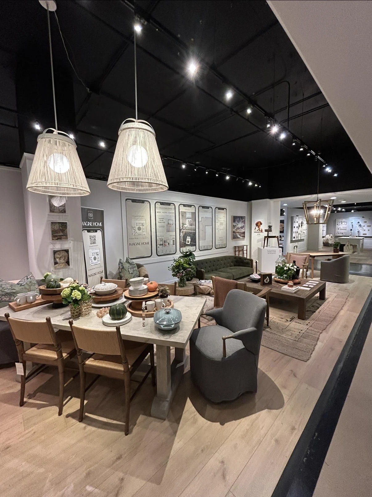 The Imagine Home booth at a design trade show, a styled dining table and living room seating area, in front of a wall of phone mockups showing the app.